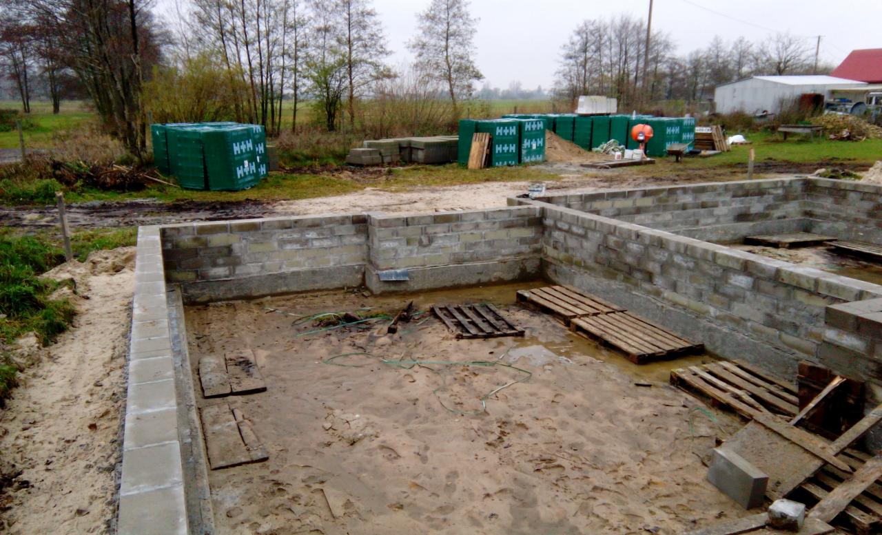 Fundamenty domu z bloczków betonowych w trakcie budowy, widoczne palety i zapasy materiałów budowlanych na zewnątrz, krajobraz wiejski w tle.