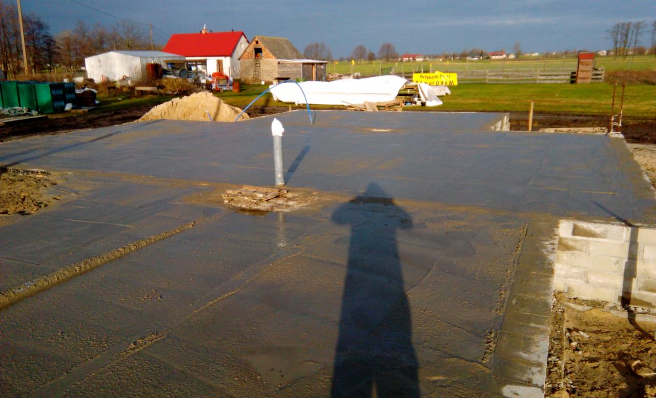 Świeżo wylana, mokra płyta fundamentowa domu jednorodzinnego na wsi, z widocznym wystającym pionowo rurociągiem i cieniem fotografa.