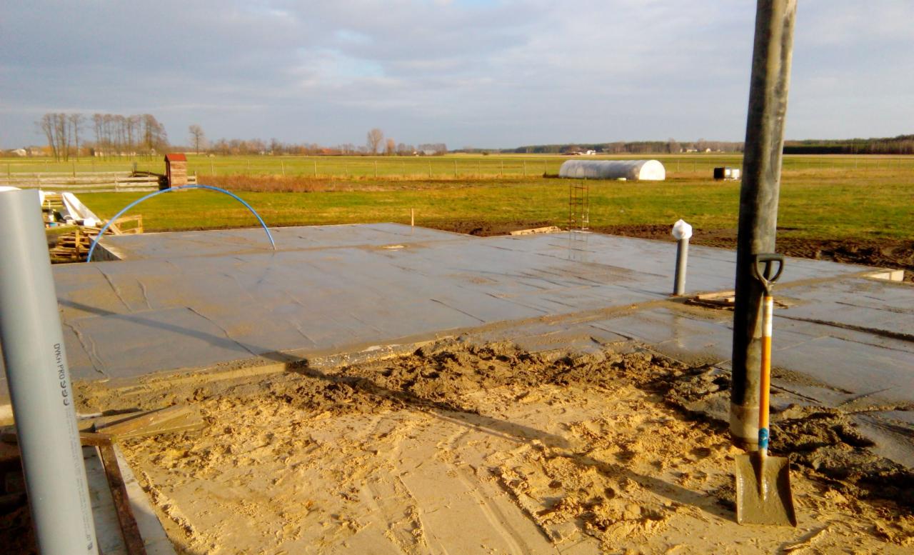 Wylana betonowa płyta fundamentowa z wystającymi rurami kanalizacyjnymi na tle łąki i szklarni, obok stoi łopata.