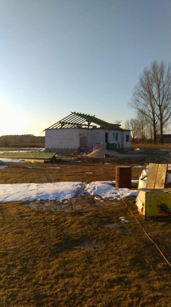 Niedokończona budowa domu jednorodzinnego z białych bloczków, widoczne drewniane elementy konstrukcji dachu, w tle pole z resztkami śniegu i drzewa.