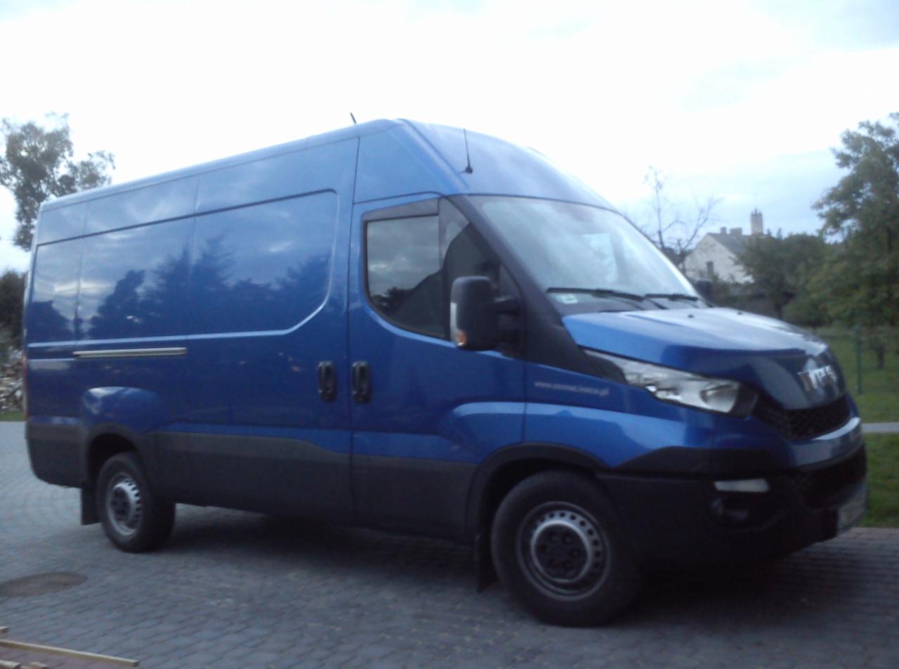 Niebieski bus dostawczy marki Iveco zaparkowany na brukowanej powierzchni, z odbiciami drzew na karoserii i budynkiem w tle.