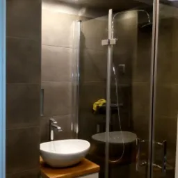 Nowoczesna łazienka z ciemnymi płytkami, kabiną prysznicową ze szklanymi drzwiami i umywalką nablatową na drewnianym blacie.