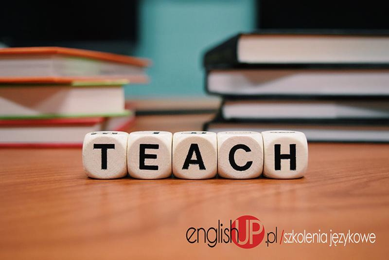Kostki z literami układające się w słowo TEACH na drewnianym biurku we Wrocławiu, w tle stosy książek i logo englishUP.pl.