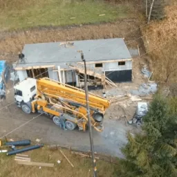 Widok z góry na plac budowy z częściowo wzniesionym budynkiem z szarym dachem, żółtą pompą do betonu oraz pracownikami w trakcie wykonywania prac.