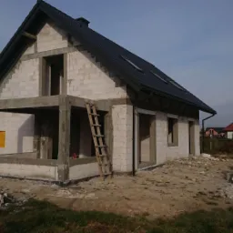 Surowy stan domu z białego bloczka, z czarnym dachem i drewnianą drabiną opartą o ścianę, widok z zewnątrz na tle innych domów.