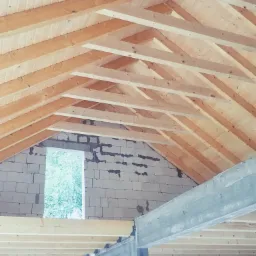 Surowy stan poddasza w trakcie budowy, widoczne drewniane krokwie i deski poszycia dachu, ściany z białego bloczka oraz betonowy słup i belka stropowa, okno z widokiem na zieleń.