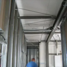 Konstrukcja ścian działowych z profili stalowych i częściowo zamontowanych płyt na suficie, widoczne przewody elektryczne i fragment sylwetki osoby w oddali.