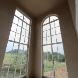 Wnętrze w stanie deweloperskim z dwoma dużymi oknami: prostokątnym i łukowym, ściany pokryte świeżym, jasnym tynkiem maszynowym, widok na zielone pola za oknem.