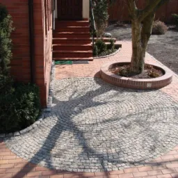 Okrągły wzór z jasnoszarej kostki ułożony na ceglanej ścieżce, otaczający drzewo z ceglanym obrzeżem, obok schody wejściowe do domu.