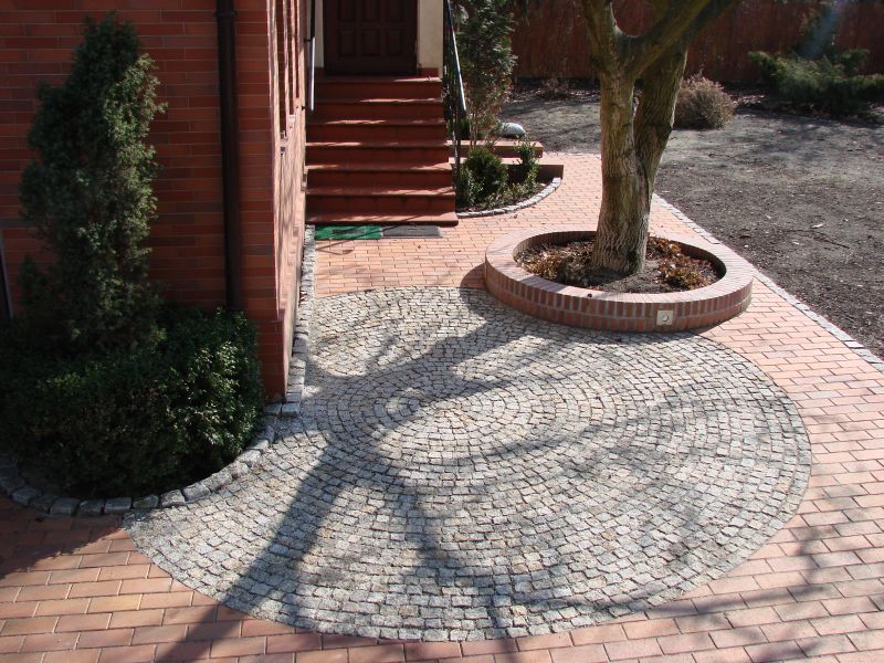 Okrągły wzór z jasnoszarej kostki ułożony na ceglanej ścieżce, otaczający drzewo z ceglanym obrzeżem, obok schody wejściowe do domu.