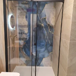 Nowoczesna kabina prysznicowa z czarnymi profilami i deszczownicą, na tle płytek w odcieniach beżu i błękitu ze złotymi akcentami. Widoczny fragment toalety.