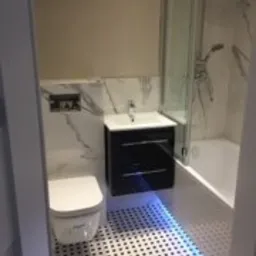 Nowoczesna łazienka z białymi płytkami imitującymi marmur i czarną szafką pod umywalką, podświetlana niebieskim światłem, widok z perspektywy drzwi.