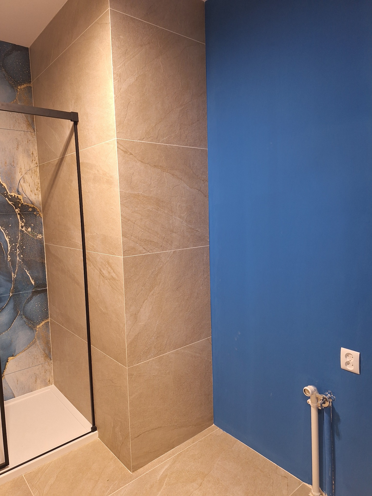 Wykończona łazienka: kabina prysznicowa z czarnymi profilami, beżowe płytki imitujące kamień i ściana pomalowana na intensywny niebieski kolor. Widoczny grzejnik.