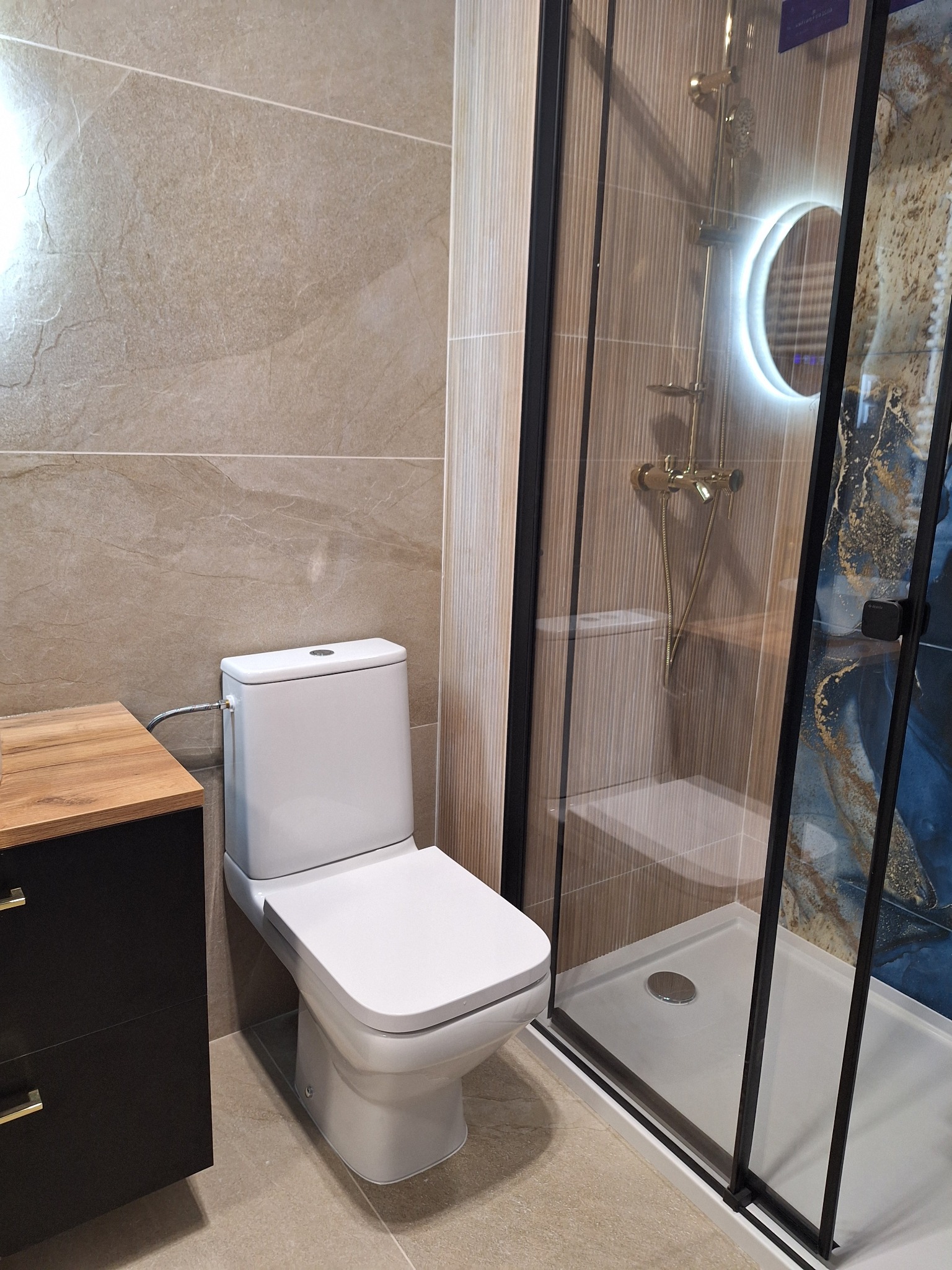 Nowoczesna łazienka po remoncie: beżowe płytki, biała toaleta, czarna szafka z drewnianym blatem i kabina prysznicowa z złotą armaturą.