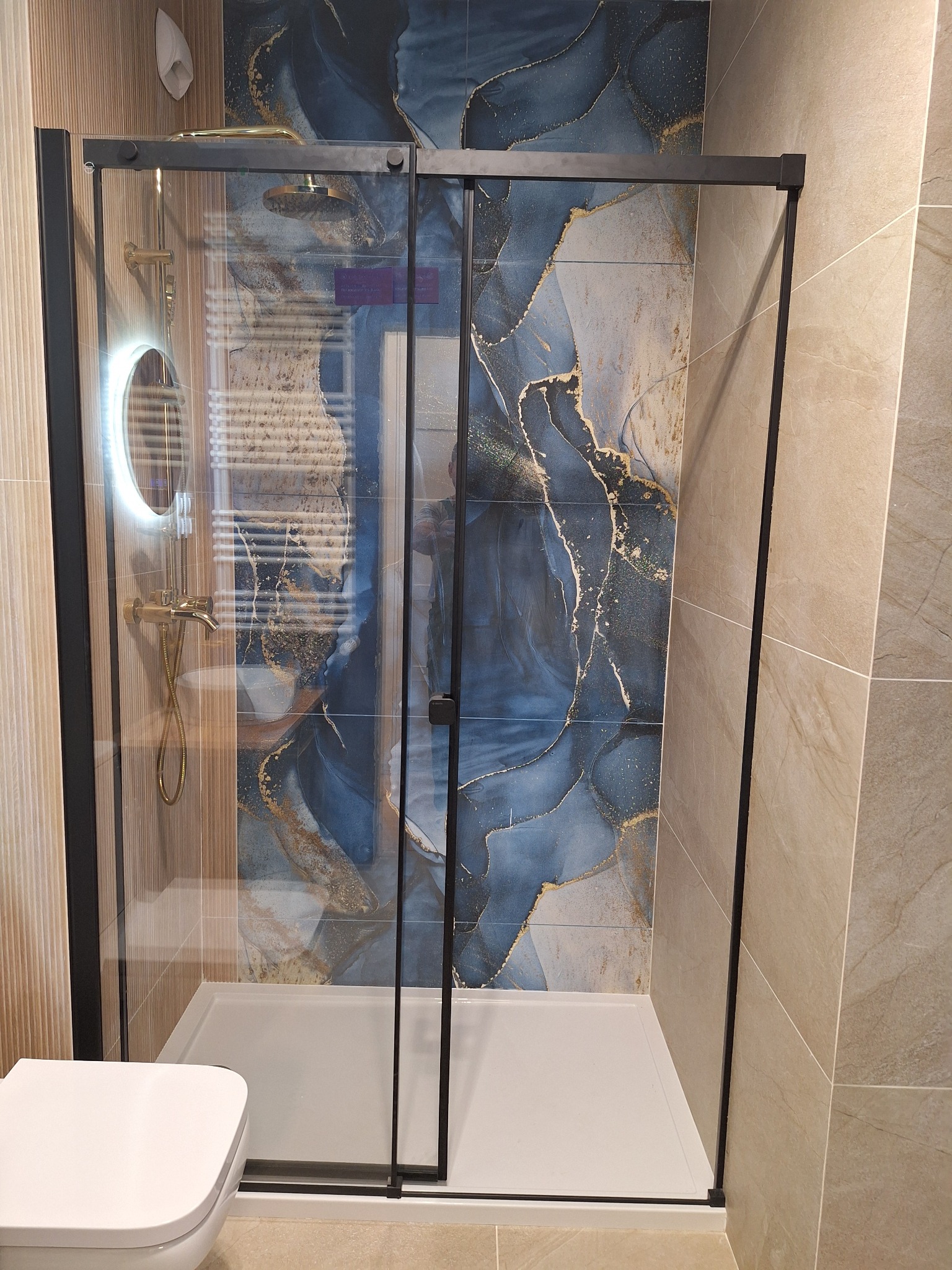 Nowoczesna kabina prysznicowa z czarnymi profilami i deszczownicą, na tle płytek w odcieniach beżu i błękitu ze złotymi akcentami. Widoczny fragment toalety.