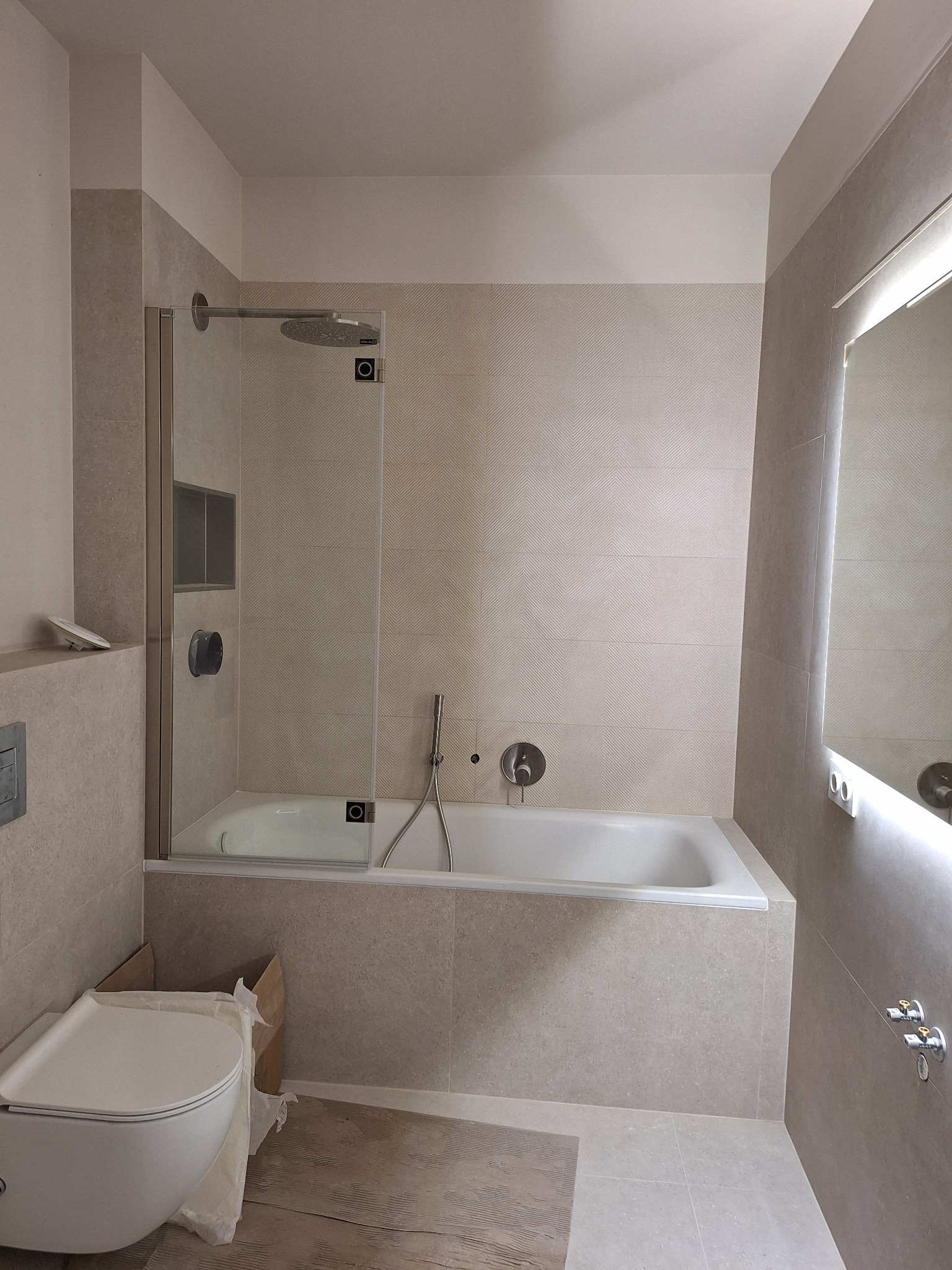 Wykończona łazienka w beżowych płytkach z kabiną prysznicową nad wanną i podświetlanym lustrem. Widoczna toaleta w trakcie montażu. Minimalistyczny design.