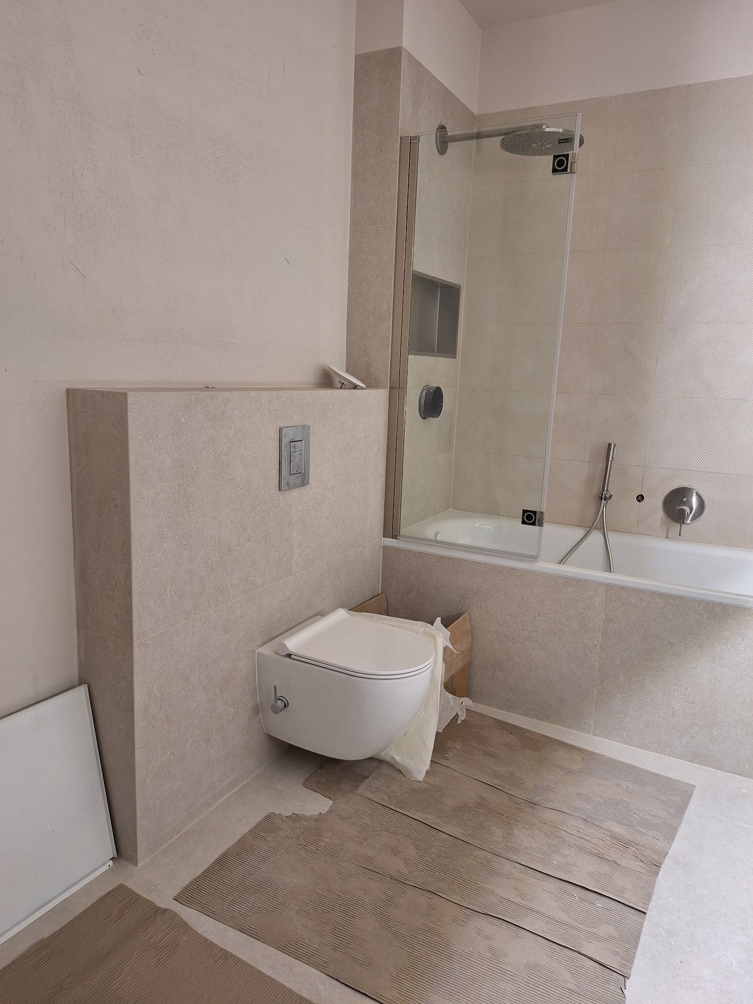 Jasna łazienka w trakcie wykańczania: podwieszana toaleta, wanna z parawanem, beżowe płytki. Podłoga zabezpieczona kartonem.