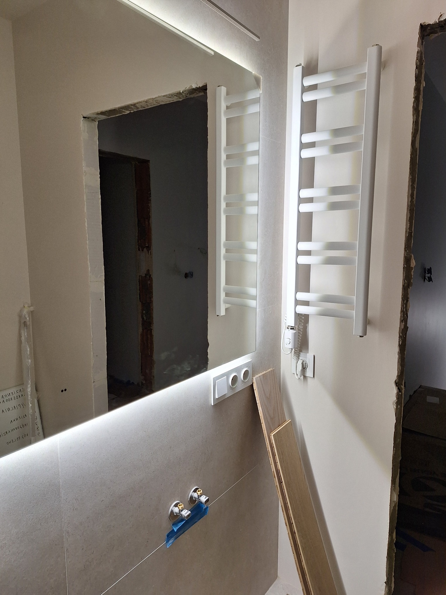 Łazienka w trakcie prac: podświetlane lustro, grzejnik drabinkowy, przygotowane przyłącza wodne. Widoczne deski podłogowe i surowe wykończenie otworu drzwiowego.