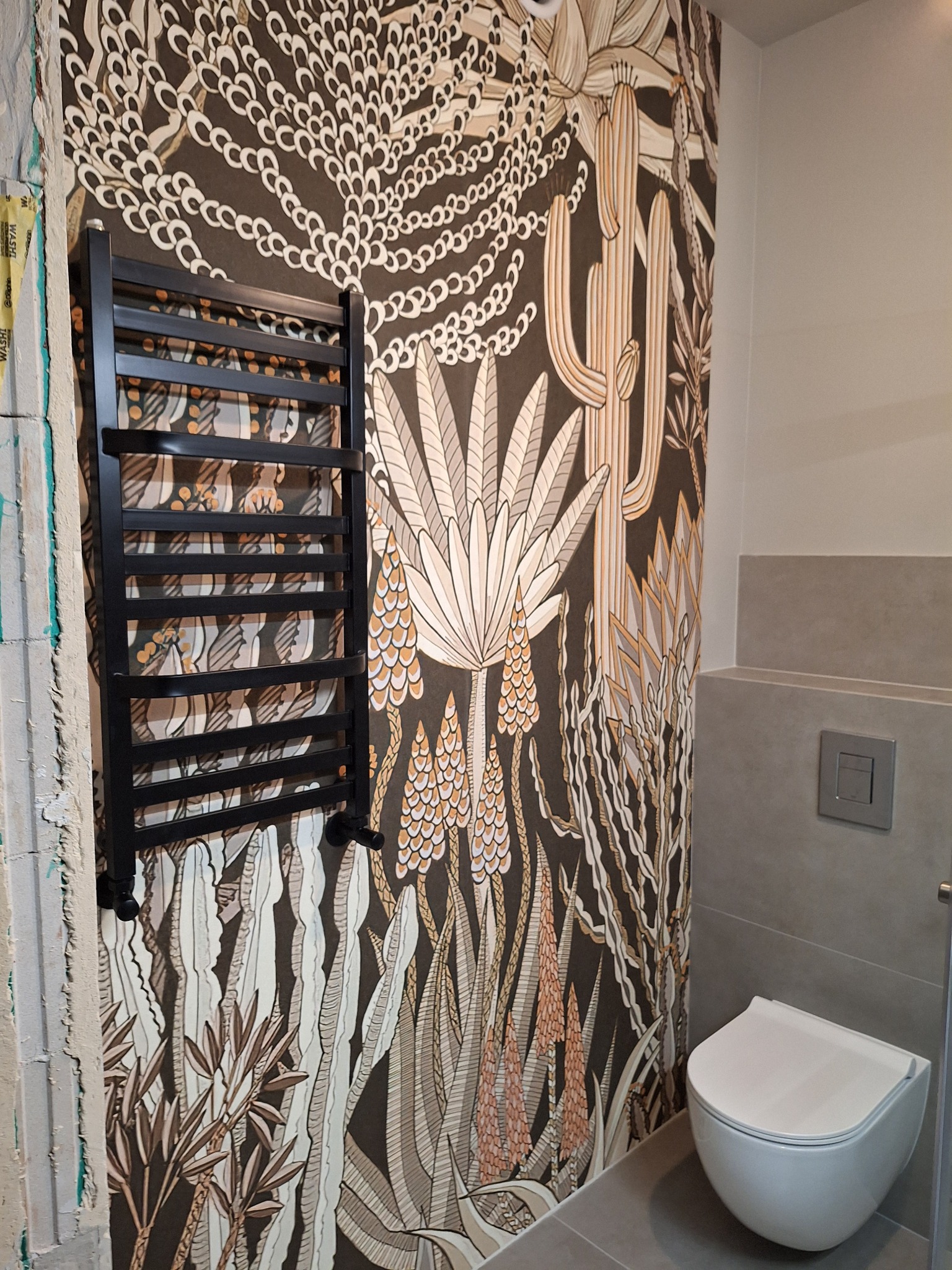 Wykończona łazienka z grzejnikiem drabinkowym na ścianie z tapetą w motywy roślinne. Widoczna toaleta i fragment ściany w szarych płytkach. Fragment ściany przed wykończeniem.