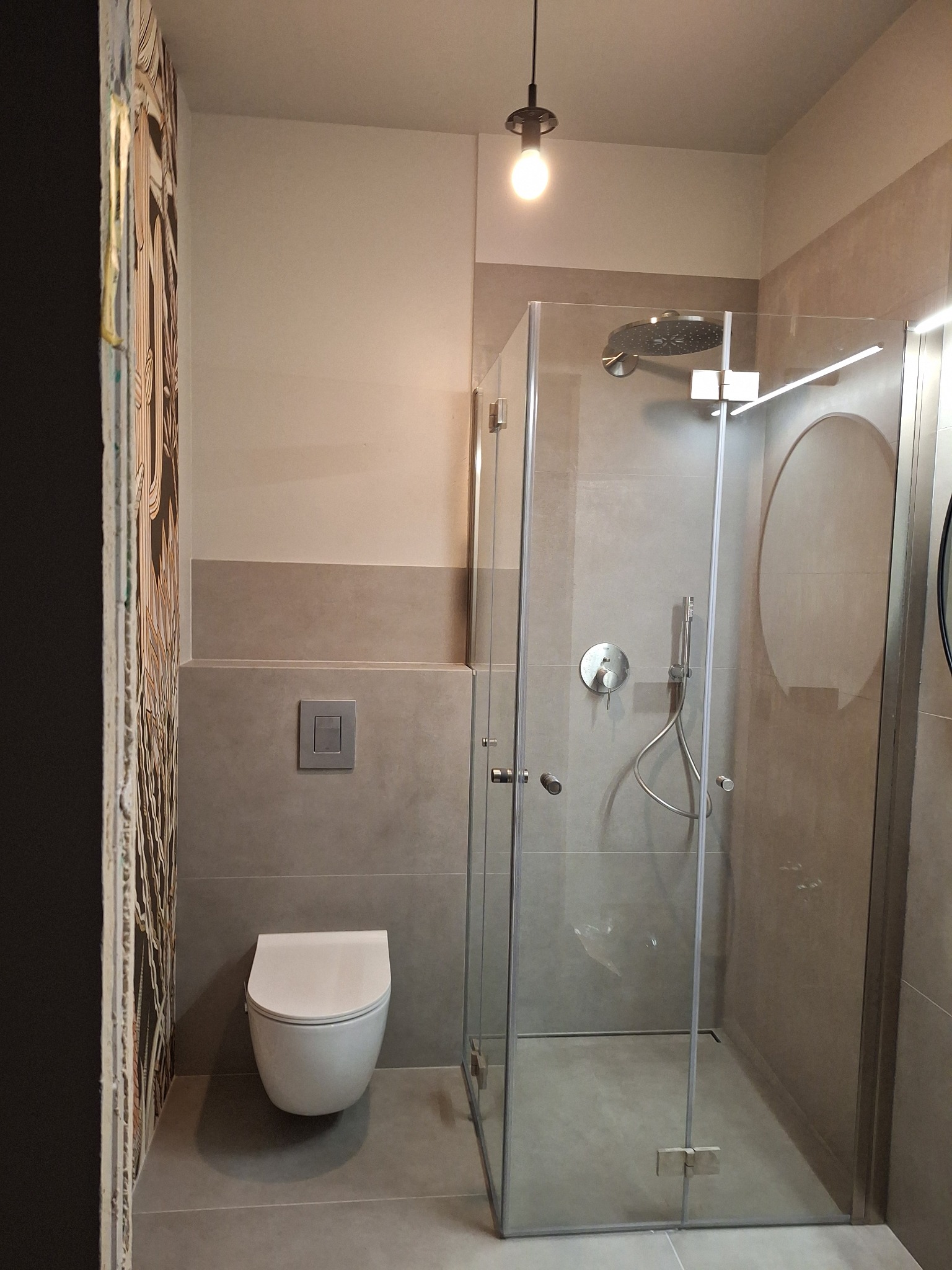 Nowoczesna łazienka w odcieniach szarości z kabiną prysznicową typu walk-in i podwieszaną toaletą. Na ścianie dekoracyjna grafika. Minimalistyczna lampa sufitowa.