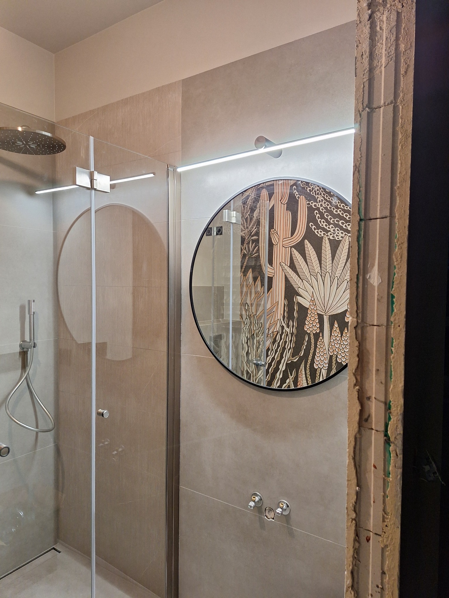 Wykończona łazienka z kabiną prysznicową, okrągłym lustrem z motywem roślinnym i oświetleniem LED. Widoczny fragment ściany w trakcie prac wykończeniowych.