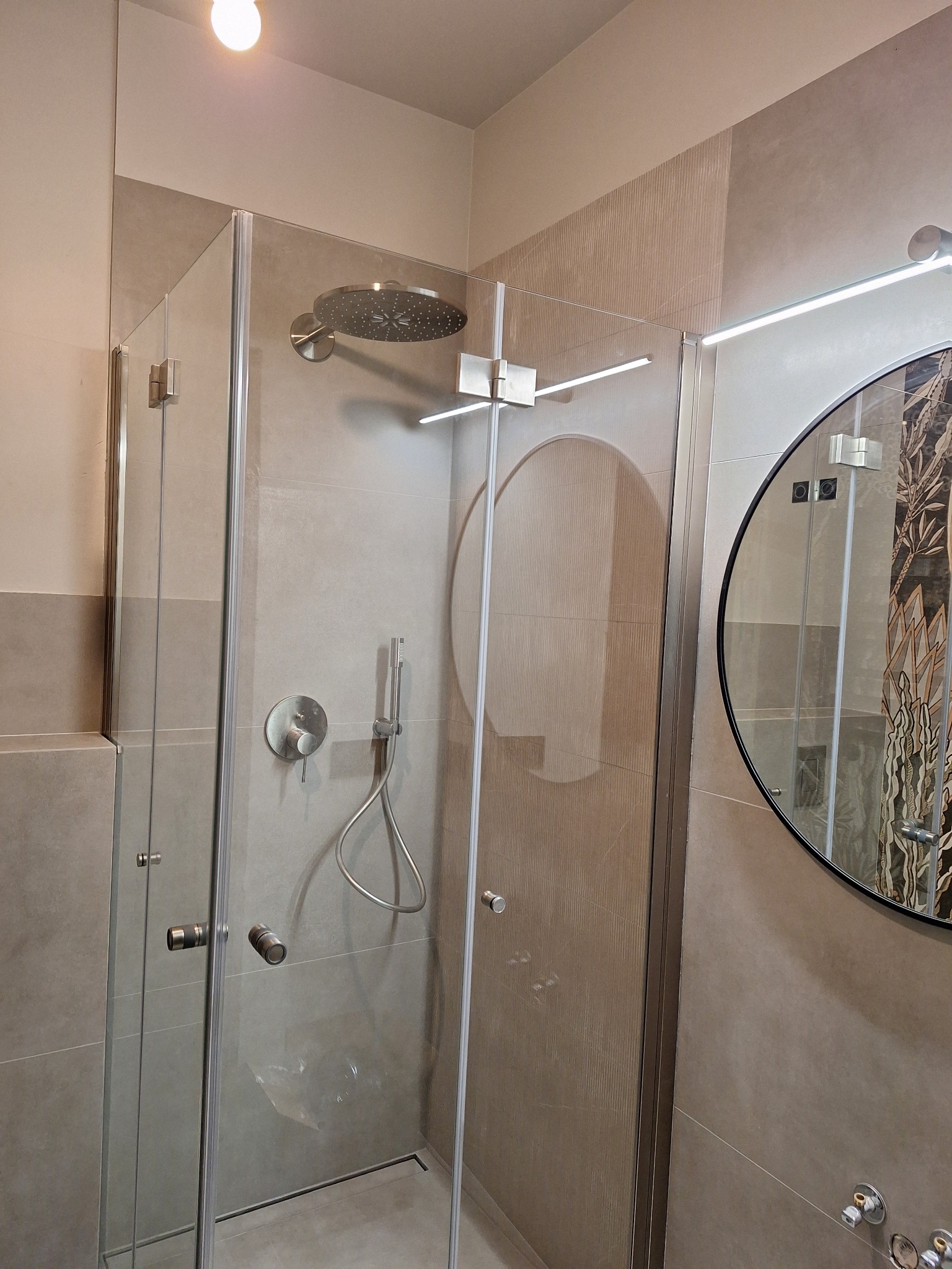 Nowoczesna kabina prysznicowa z deszczownicą i okrągłym lustrem w wykończonej łazience. Beżowe płytki z subtelnym wzorem. Minimalistyczny design.