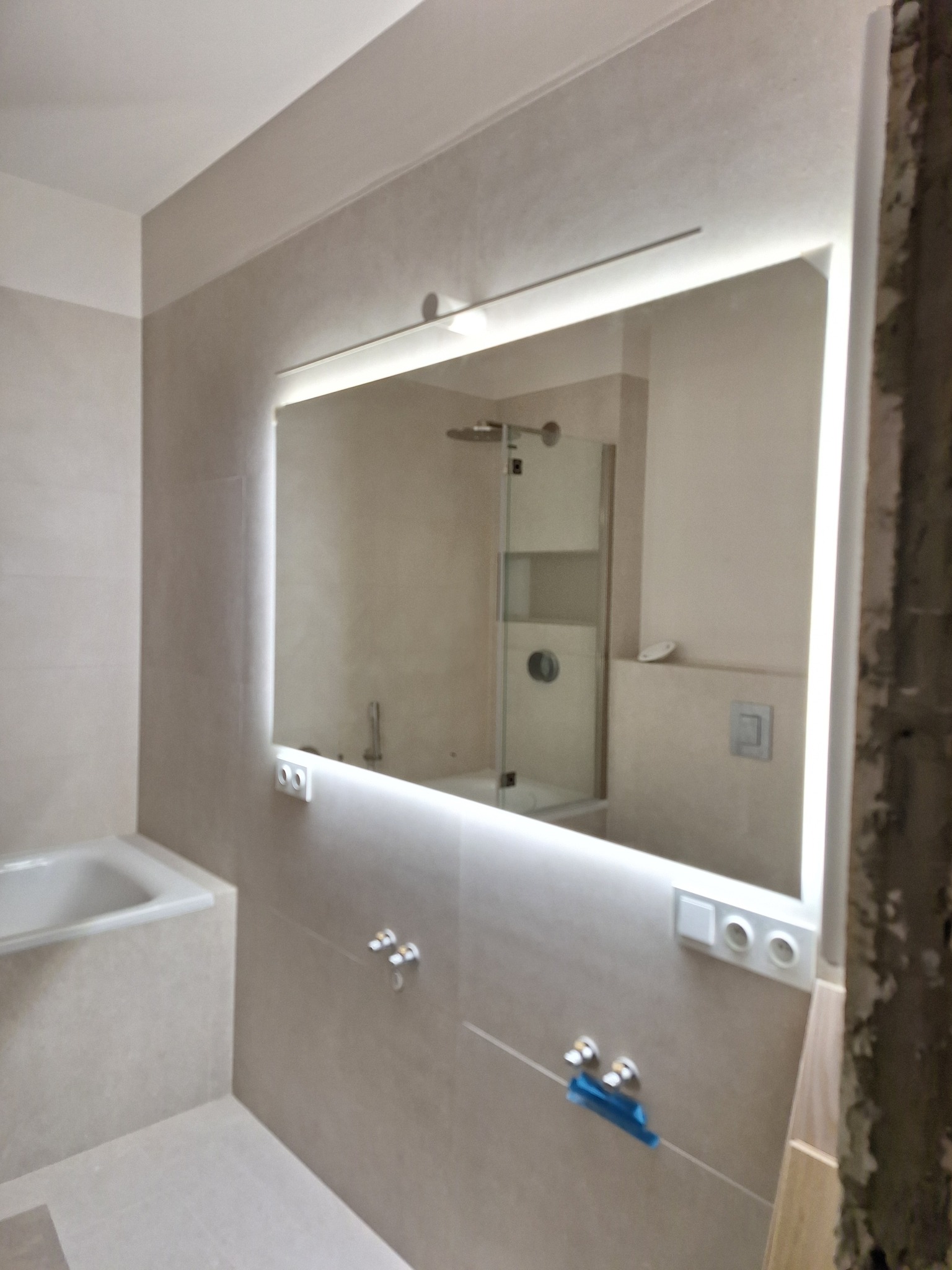 Nowoczesna łazienka z podświetlanym lustrem i beżowymi płytkami. Widoczne bateria, wanna i fragment kabiny prysznicowej. Częściowo wykończone wnętrze.