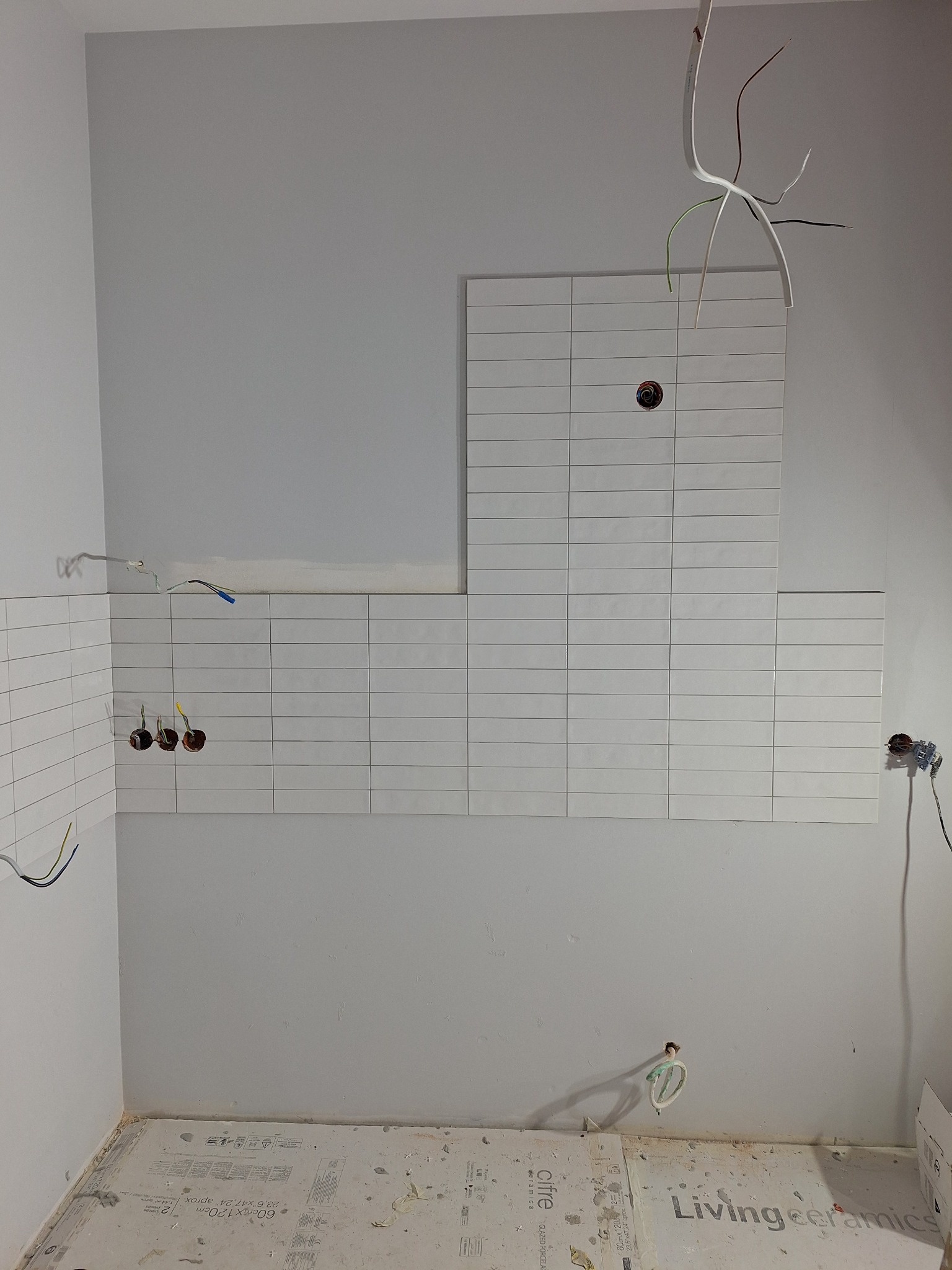 Ściana w trakcie układania białych, prostokątnych płytek. Widoczne wystające kable elektryczne i gniazdka. Podłoga zabezpieczona kartonem.