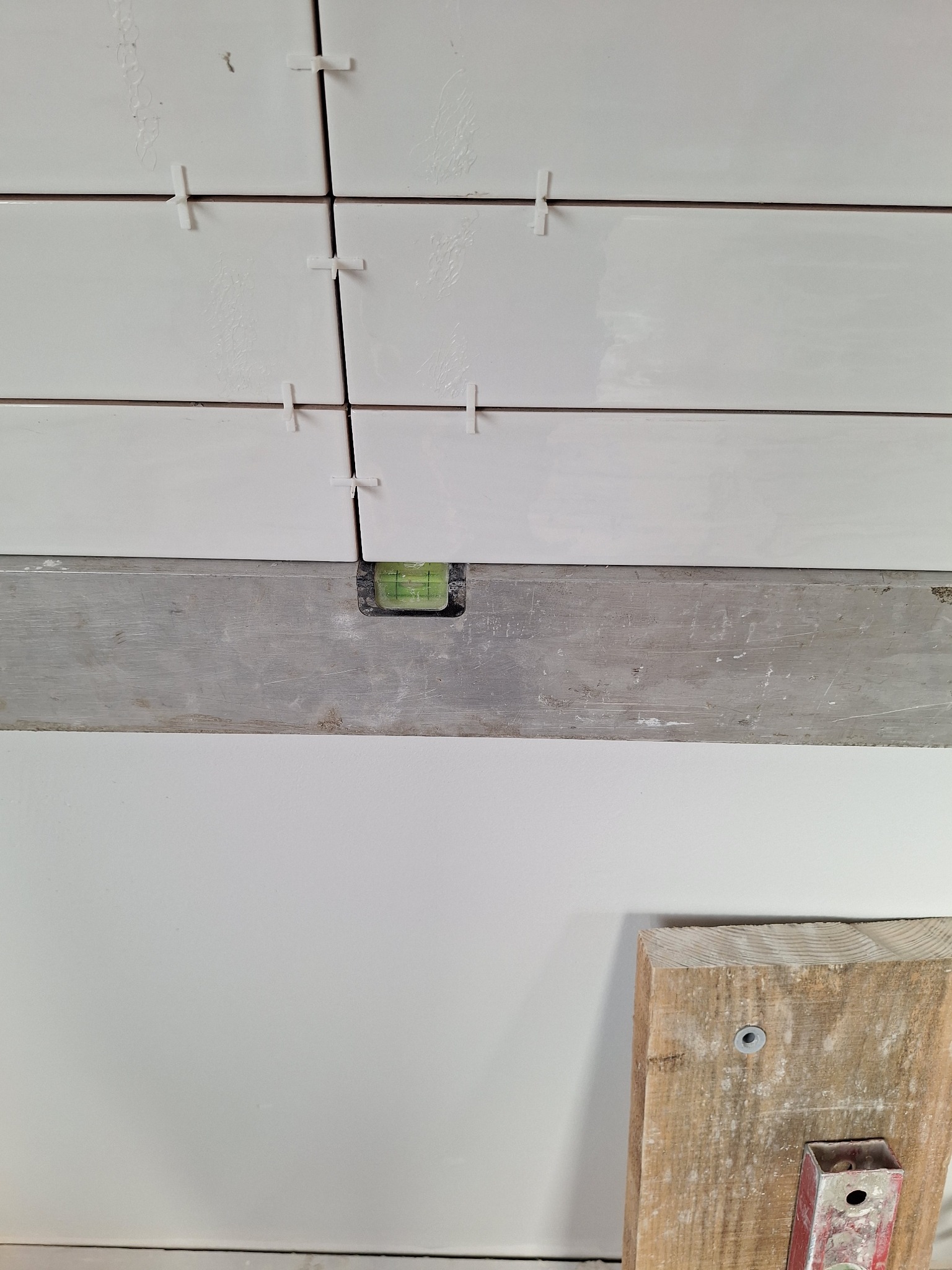 Świeżo ułożone, białe, prostokątne płytki z krzyżykami dystansowymi, poziomowane przy użyciu poziomicy. Widoczny fragment drewnianej podpory.