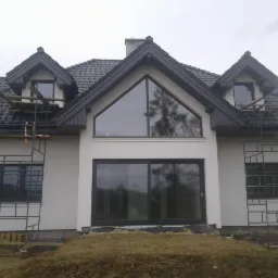 Nowoczesny dom jednorodzinny z dużymi oknami, świeżo otynkowaną elewacją i rozstawionymi rusztowaniami, teren wokół budynku jeszcze niezagospodarowany.