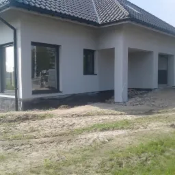 Nowoczesny dom jednorodzinny z jasną elewacją i czarnymi ramami okiennymi, widoczna izolacja fundamentów i teren wokół budynku w trakcie prac wykończeniowych.