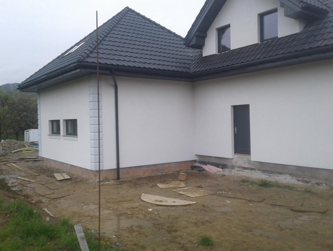 Nowoczesny dom jednorodzinny z grafitową dachówką i jasną elewacją, widoczne wykończenia narożników, teren wokół domu w trakcie prac ziemnych.