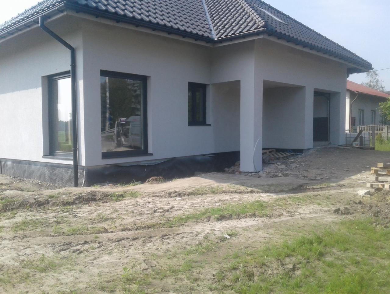 Nowoczesny dom jednorodzinny z jasną elewacją i czarnymi ramami okiennymi, widoczna izolacja fundamentów i teren wokół budynku w trakcie prac wykończeniowych.