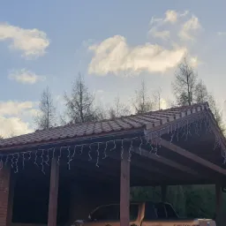 Zadaszenie garażowe z brązową konstrukcją drewnianą i czerwoną dachówką, ozdobione białymi lampkami zwisającymi z okapu, częściowo zasłaniające zaparkowany samochód, na tle błękitnego nieba...