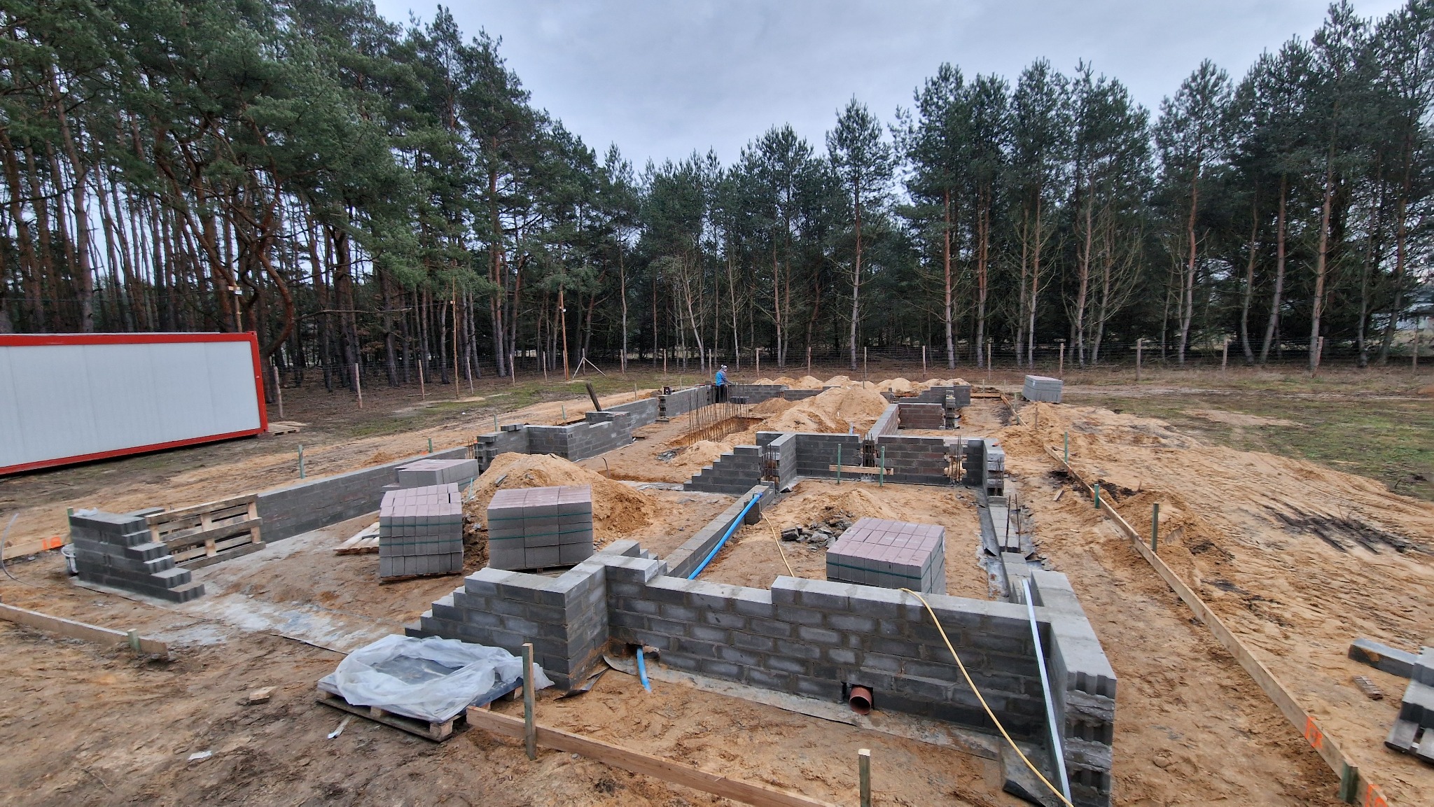 Fundamenty domu z bloczków betonowych na piaszczystym podłożu, widoczne zbrojenia, w tle las i kontener budowlany.