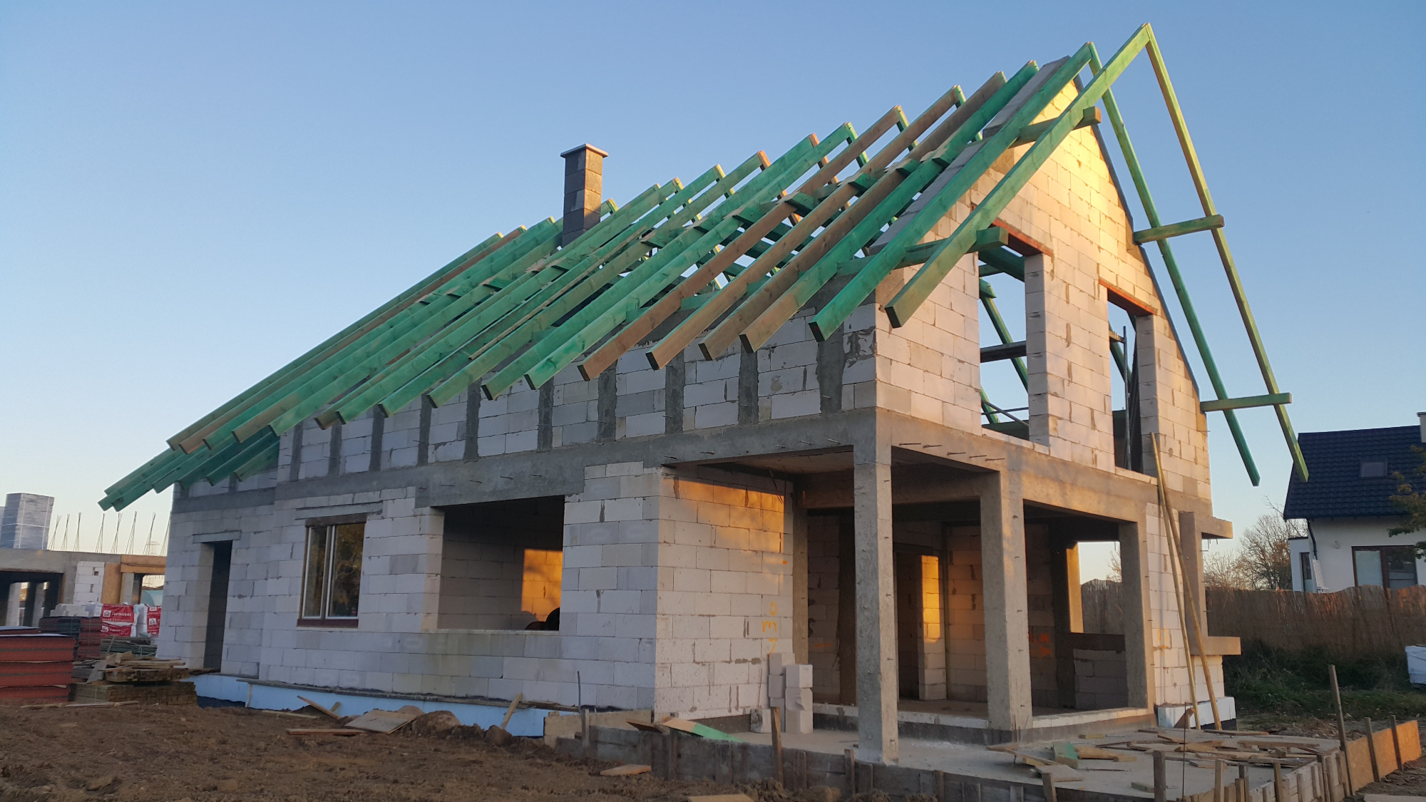 Surowy stan domu z białego betonu komórkowego, z zielonymi elementami więźby dachowej i kominem z cegły, w świetle wschodzącego słońca.