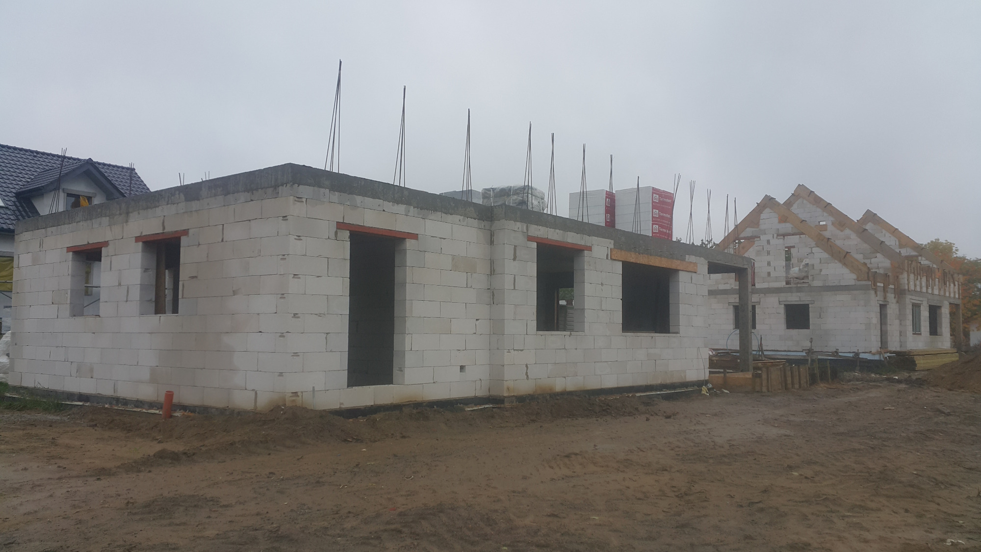 Budowa domu jednorodzinnego z betonu komórkowego w stanie surowym otwartym; widoczne ściany, otwory okienne z nadprożami, strop oraz więźba dachowa drugiego budynku w tle, teren budowy po deszczu.