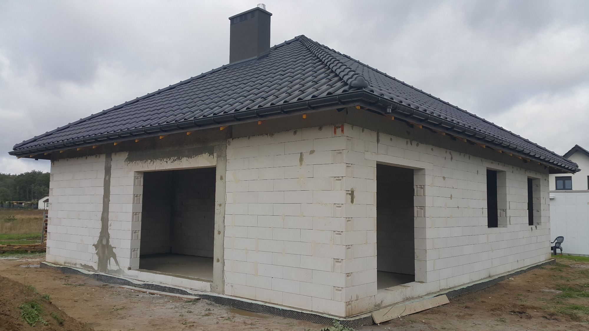 Surowy stan domu jednorodzinnego z białych bloczków, czarny dach z kominem, widoczne otwory okienne i drzwiowe, teren wokół w trakcie przygotowania.