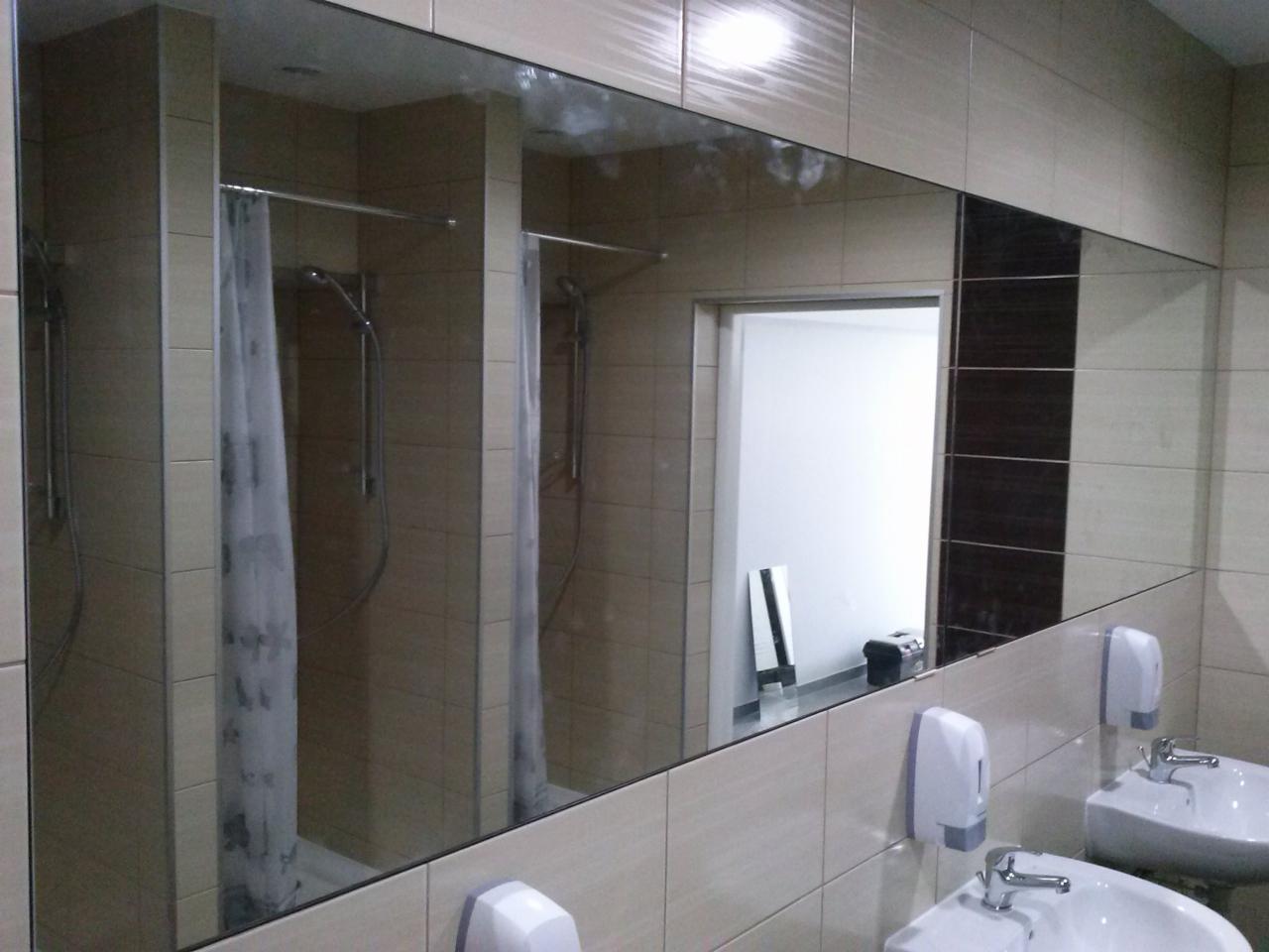 Duże lustro ścienne w publicznej łazience odbija kabiny prysznicowe z zasłonami, umywalki z dozownikami mydła i białe płytki na ścianach.