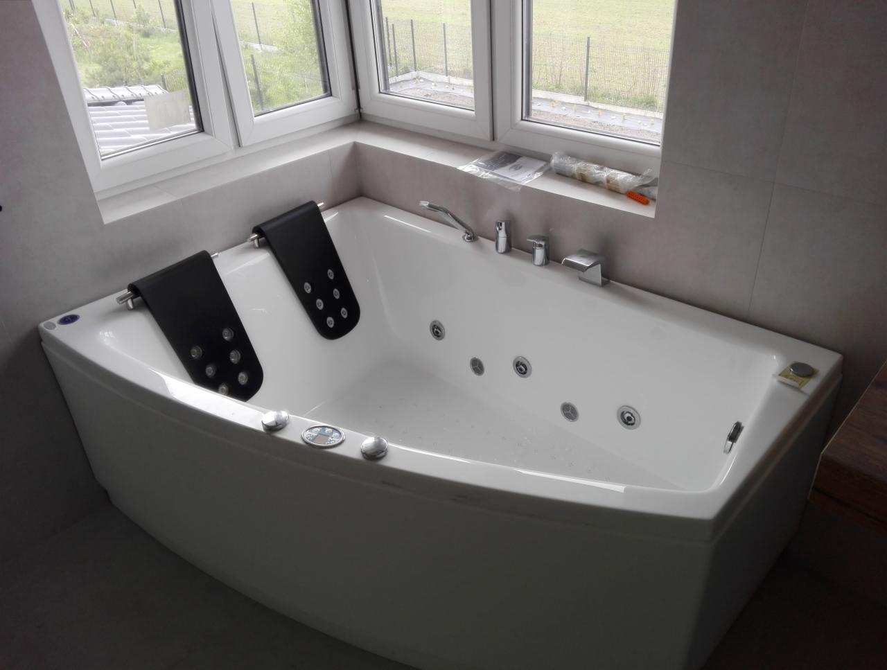 Biała, narożna wanna z hydromasażem i czarnymi zagłówkami pod oknem, świeżo wykończona, z widocznymi szarymi płytkami na ścianach.