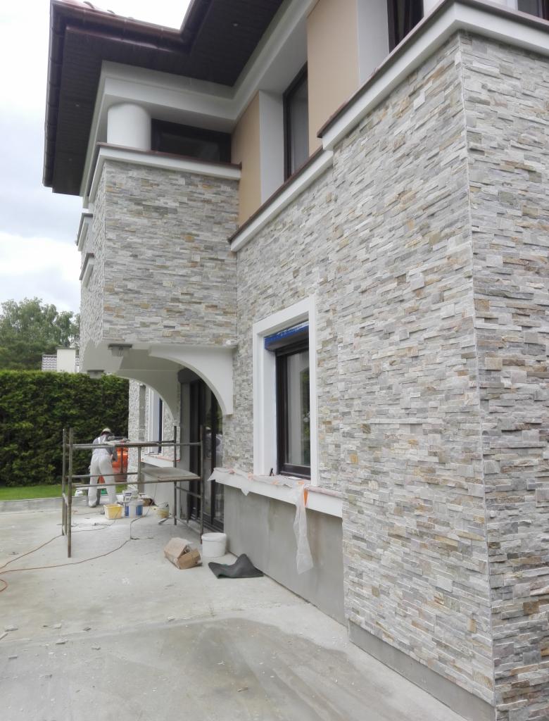 Układanie kamienia elewacyjnego na ścianie domu jednorodzinnego, widoczne rusztowanie i pracownik w kombinezonie ochronnym, ściana w trakcie prac wykończeniowych.