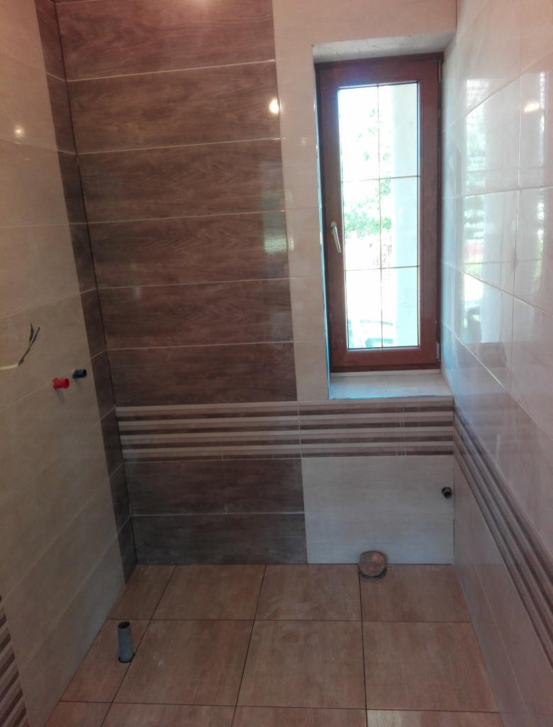 Wykończona łazienka z brązowo-beżowymi płytkami, drewnianym oknem i widocznymi przyłączami wodnymi.