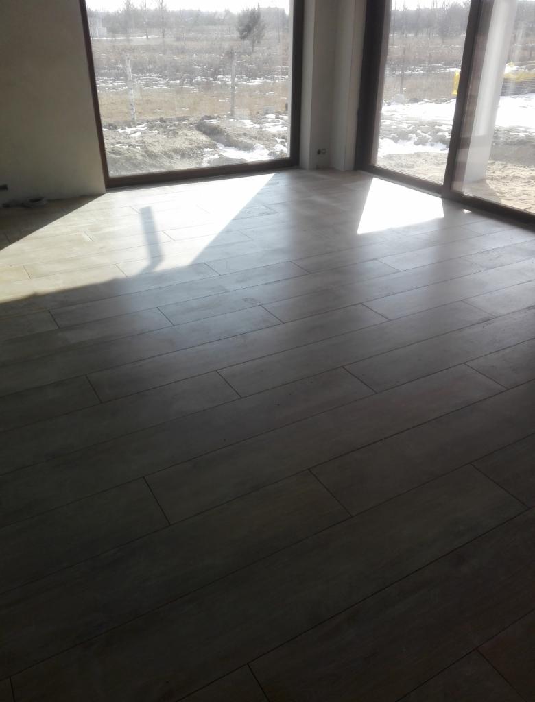 Ułożona podłoga z płytek imitujących drewno w jasnym odcieniu, widok z niskiego kąta, z dużymi oknami w tle i silnym światłem słonecznym padającym na podłogę, tworzącym kontrastowe cienie.