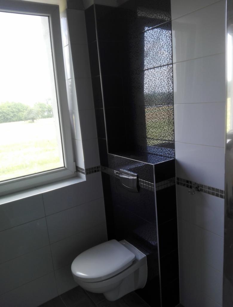 Nowoczesna łazienka z wiszącą toaletą, czarno-białe płytki z mozaikowym akcentem, okno z widokiem na zieleń, minimalistyczny design.