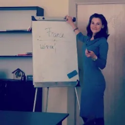 Nauczycielka w niebieskiej sukience pisze markerem na białej tablicy słowa 'France' i 'cuisine' podczas lekcji francuskiego.