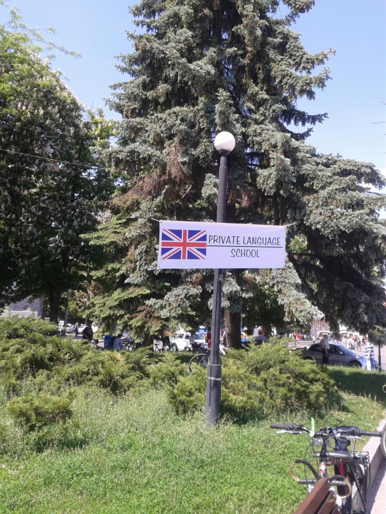 Baner z flagą Wielkiej Brytanii i napisem 'Private Language School' umieszczony na słupie oświetleniowym w parku, rowery na pierwszym planie.
