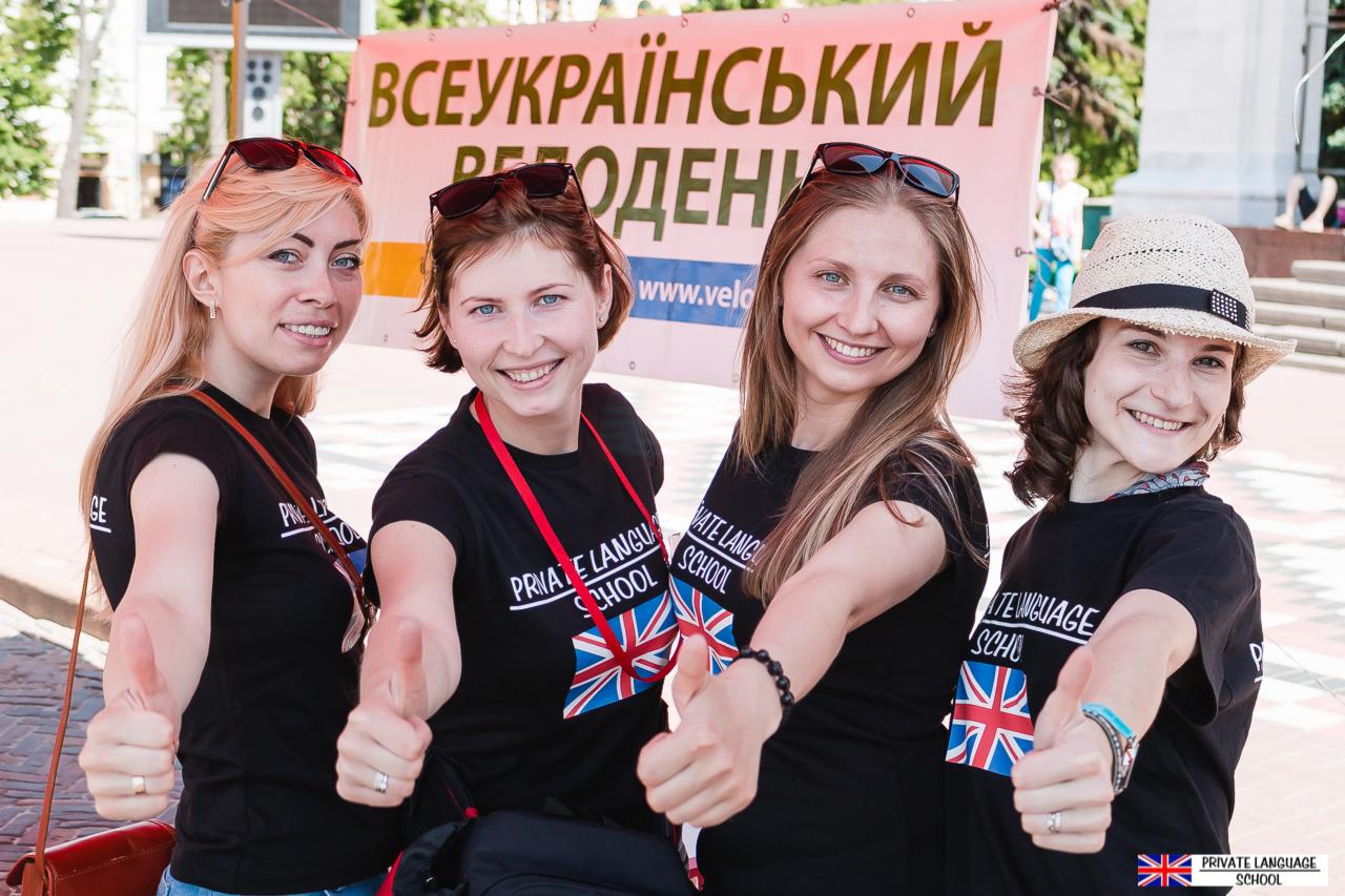 Cztery uśmiechnięte kobiety w czarnych koszulkach z logo szkoły językowej na tle baneru reklamowego, pokazujące kciuki w górę.