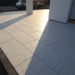 Nowo ułożona, jasnoszara kostka brukowa na posesji, z widocznym obrzeżem z ciemniejszych kostek i świeżo przygotowanym gruntem pod dalsze prace, oświetlona popołudniowym słońcem.