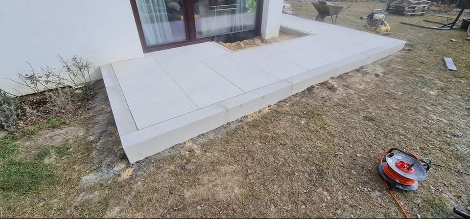 Wykończony taras z jasnych, prostokątnych płyt betonowych przylegający do budynku, z widoczną niezabudowaną przestrzenią przy drzwiach tarasowych i zwiniętym kablem elektrycznym w trawie obok.