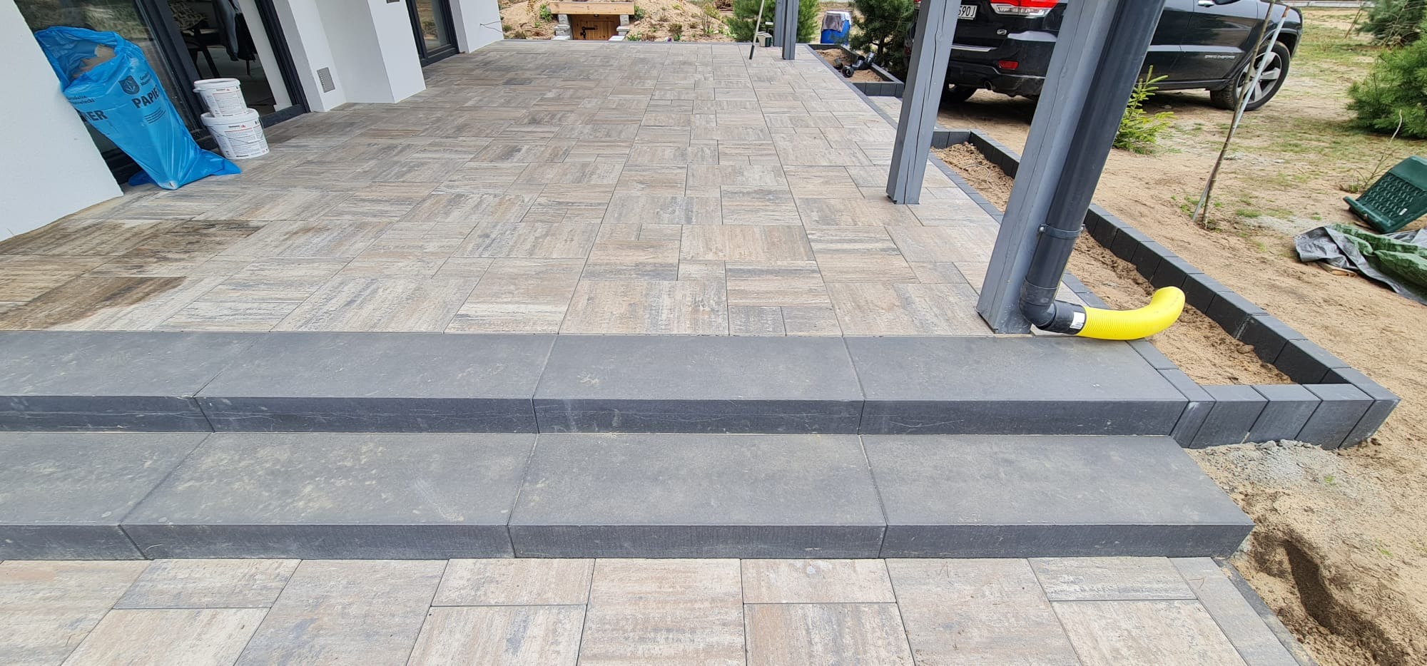 Ułożona kostka brukowa na tarasie z dwoma stopniami, widoczna rura spustowa z żółtą końcówką, worki i puszki z farbą przy ścianie domu.